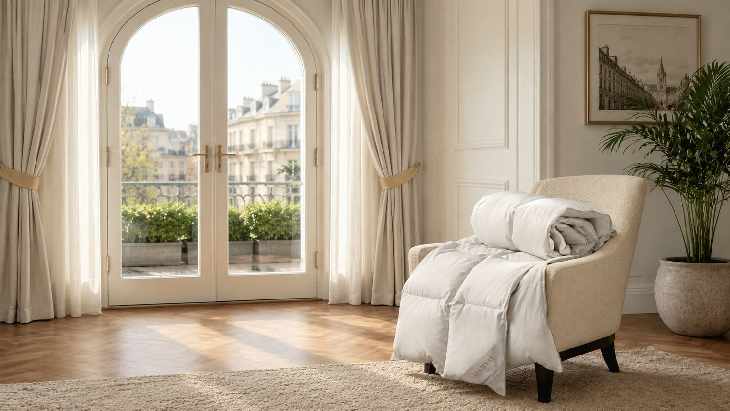 A cozy cream-colored armchair in a bright, elegant room, draped with a matching folded quilted goose down comforter. The room features tall beige curtains framing a large arched French door with a Parisian view, a wooden floor, a beige rug, and a potted plant.