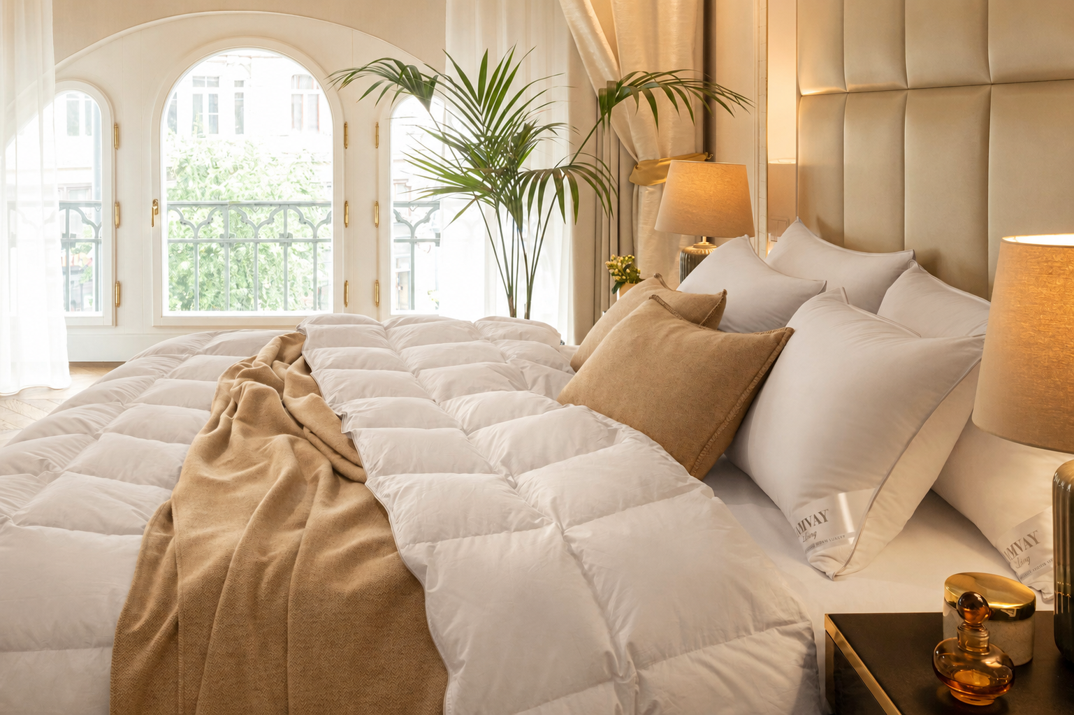 A neatly made bed in a bright, elegant bedroom featuring a white quilted comforter, white pillows, tan accent pillows and throw blanket, a tufted beige headboard, bedside lamps, and large arched windows in the background.