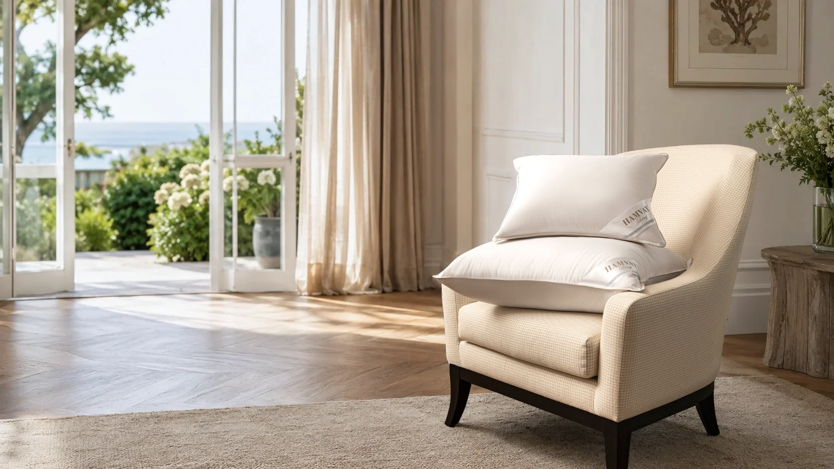 A cream upholstered armchair with dark wooden legs sits in a bright, modern living room with large windows, beige curtains, and a view of the sea. The chair is styled with two white pillows.