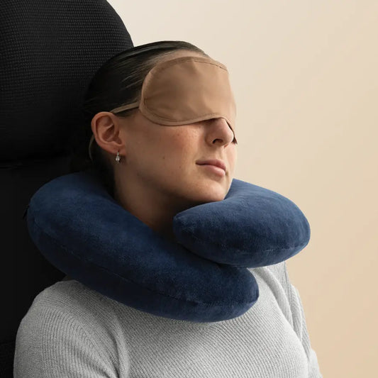 Woman using a navy blue travel neck pillow with a beige eye mask.